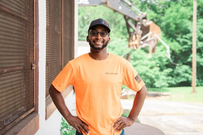 Clinton Harris III in a CJ's Ventures shirt standing in front of demolition equipment on a job site.